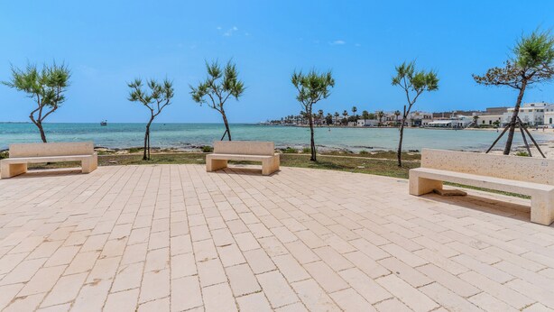 A paved walkway with benches and trees, leading to a beach with a clear view of the sea and distant buildings. - 4000 Casa Chiaraluna by Barbarhouse (Porto Cesareo)