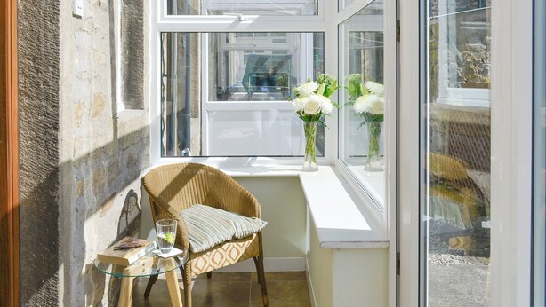 Une pièce d’angle avec un fauteuil en osier, une petite table et une fenêtre ornée de fleurs blanches.
