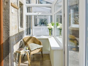 Une pièce d’angle avec un fauteuil en osier, une petite table et une fenêtre ornée de fleurs blanches.