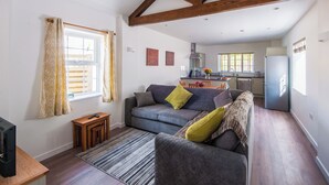 A modern living room with a sofa, a small table, and a TV. There is a kitchen in the background with a refrigerator and a window. - Beech Cottage - UK11845 (Carkeel, near Saltash)