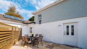 A patio with a table and chairs, a wooden fence, a white building with a grey roof, and solar panels on the roof. - Beech Cottage - UK11845 (Carkeel, near Saltash)