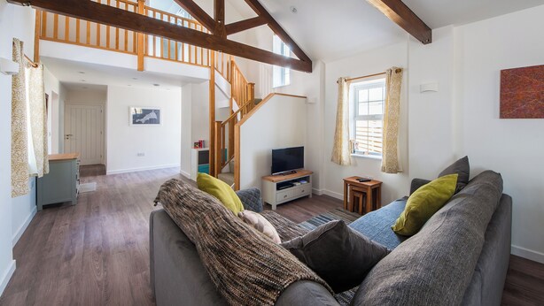 A spacious living room with a high ceiling, exposed wooden beams, a staircase, a sofa set, and a television. - Beech Cottage - UK11845 (Carkeel, near Saltash)