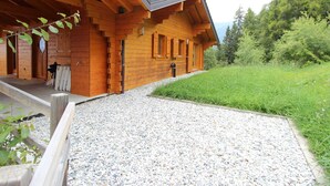 Une cabane en bois avec un toit en pente, un chemin de gravier menant à l’entrée et un paysage verdoyant luxuriant en arrière-plan.