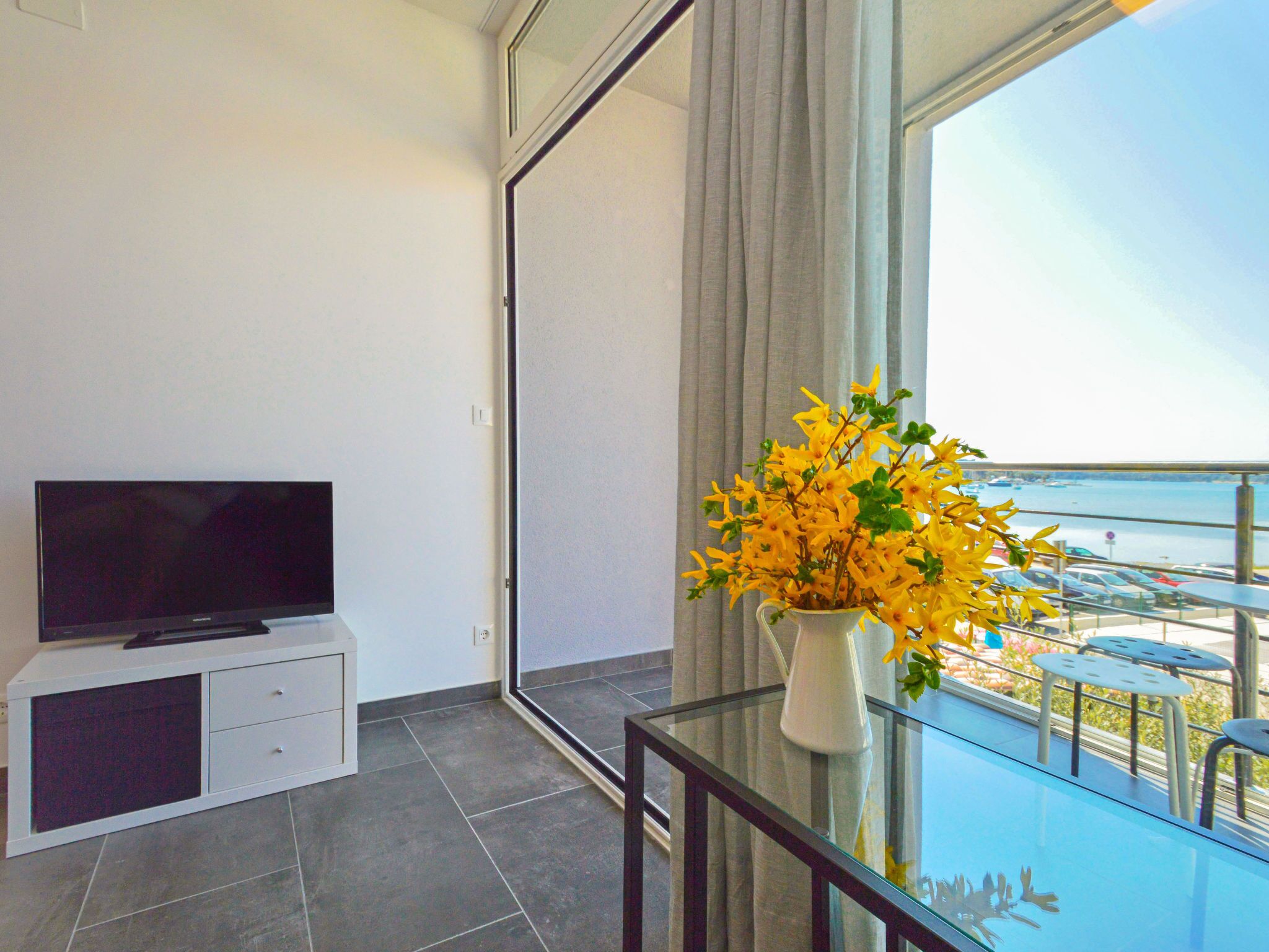 A modern room with a flat-screen TV on a white cabinet, a glass table with a vase of yellow flowers, and a balcony with a view of the sea and parked cars.