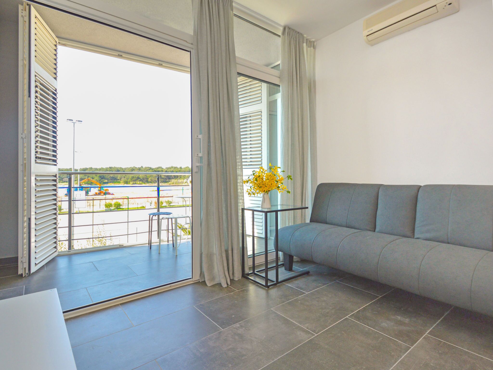 A modern living room with a grey sofa, a small table with a vase of yellow flowers, and a view of a construction site through large windows.