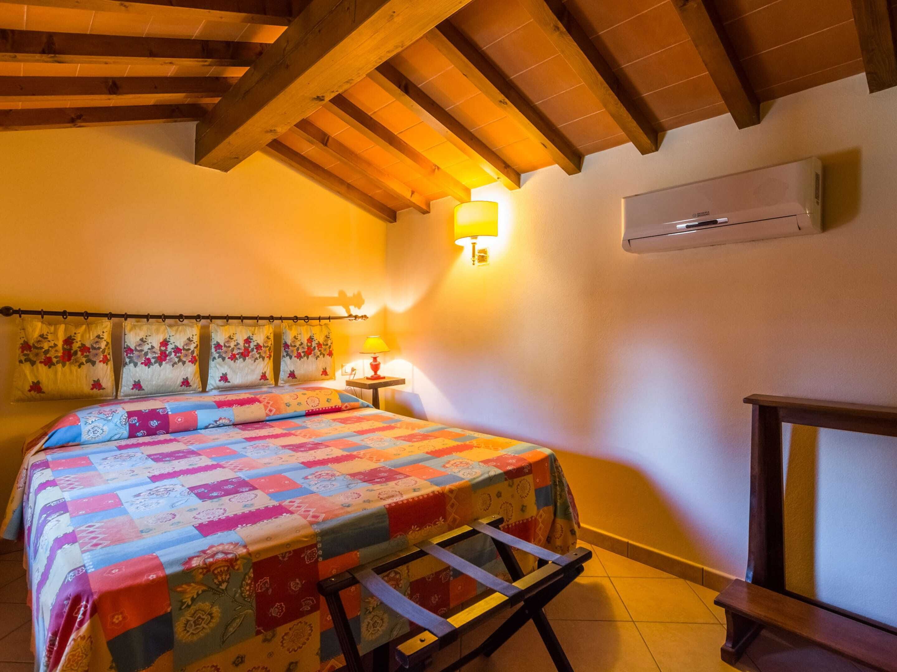 Un dormitorio con techo de madera, una cama con colcha de colores, una mesita de noche con lámpara y un equipo de aire acondicionado en la pared.
