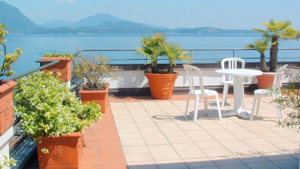 A terrace with potted plants, white chairs, and a view of a lake and mountains. - Cozy Mansion near Lake in Baveno Italy (Baveno)
