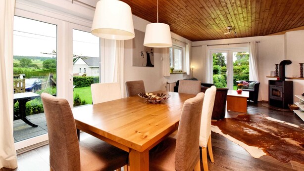 Een eetgedeelte met een houten tafel en stoelen, grote ramen en uitzicht op de omgeving.