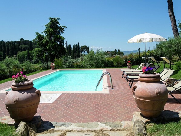 Une piscine rectangulaire avec une terrasse carrelée, entourée de chaises longues et d’un parasol.
