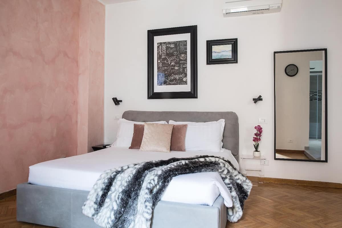 A bedroom with a bed, a bedside table with a vase of flowers, a framed picture on the wall, and a mirror.