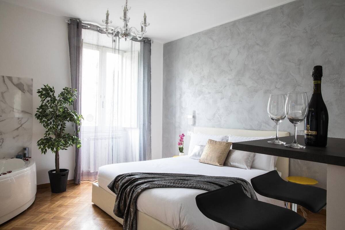 A modern bedroom with a bed, a potted plant, a chandelier, a bottle of wine, and two glasses on a nightstand.