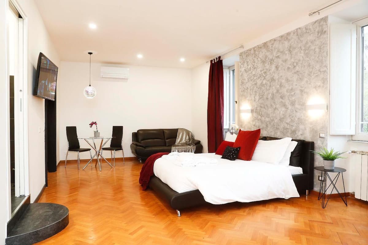 A hotel room with a bed, a sofa, a dining table with chairs, a TV, and a window with curtains.