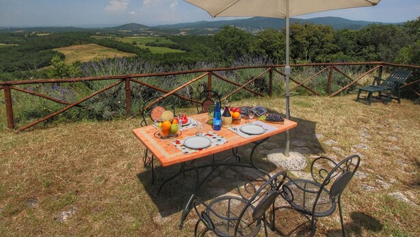 An outdoor dining area with a table set for two, surrounded by chairs, a table umbrella, and a scenic view of rolling hills and vineyards. - Belvilla by OYO Olmi (Casole d'Elsa)
