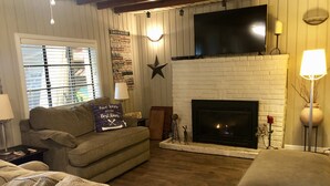 A cozy living room with a fireplace, a television mounted above it, and a couch with a decorative pillow. - Raper’s Hollow Waterfront Cabin (Eucha)