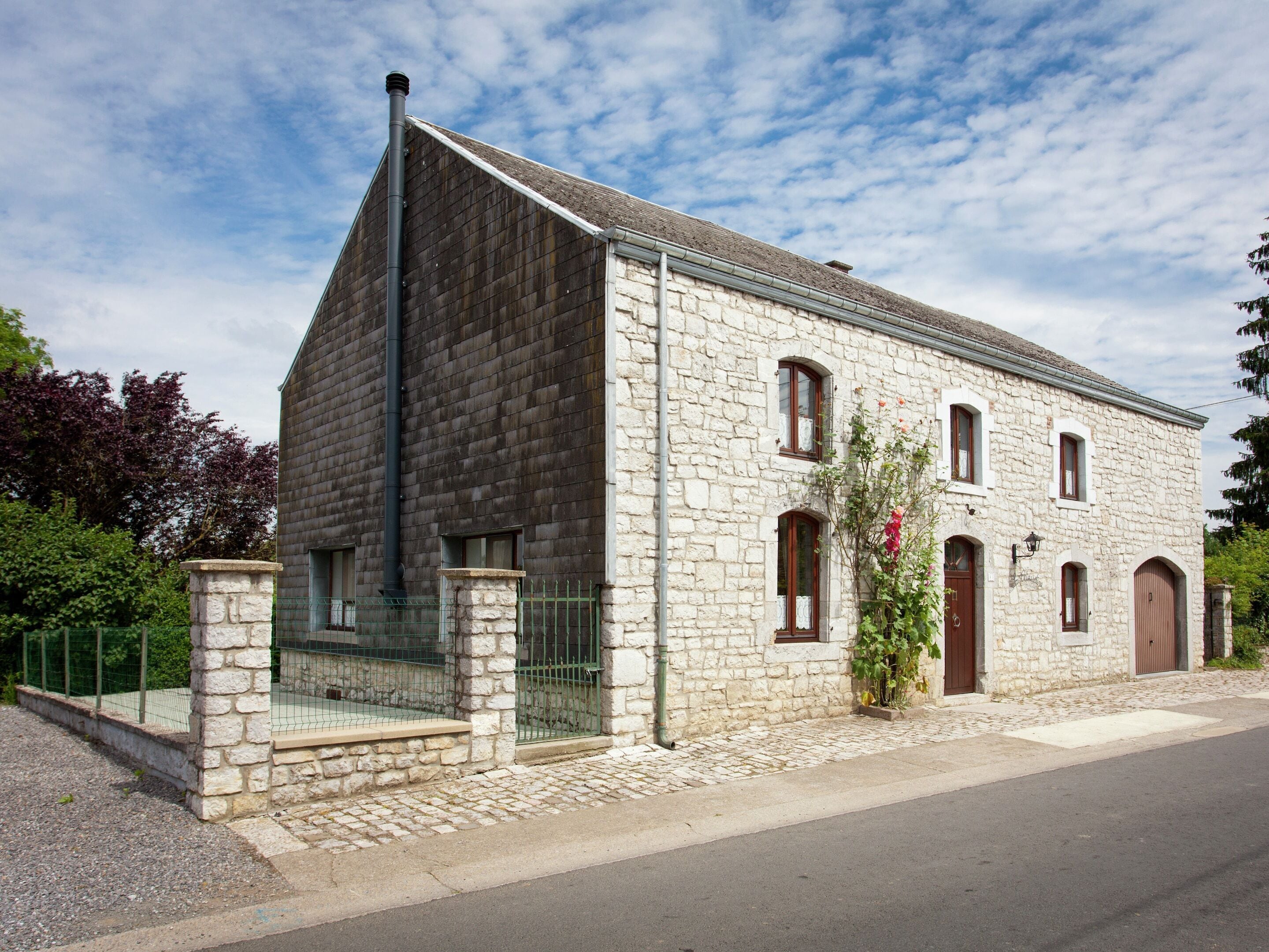Un bâtiment en pierre avec un toit en pente, une cheminée et un petit jardin agrémenté d’une plante à fleurs.
