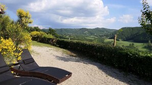 Ein Kiesweg führt zu einer Sitzgelegenheit mit Korbsesseln und Blick auf grüne Hügel und einen klaren Himmel.