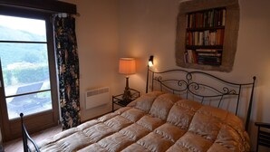 Ein Schlafzimmer mit einem großen Bett, einem Nachttisch mit Lampe, einem Bücherregal und einem Fenster mit Vorhängen.