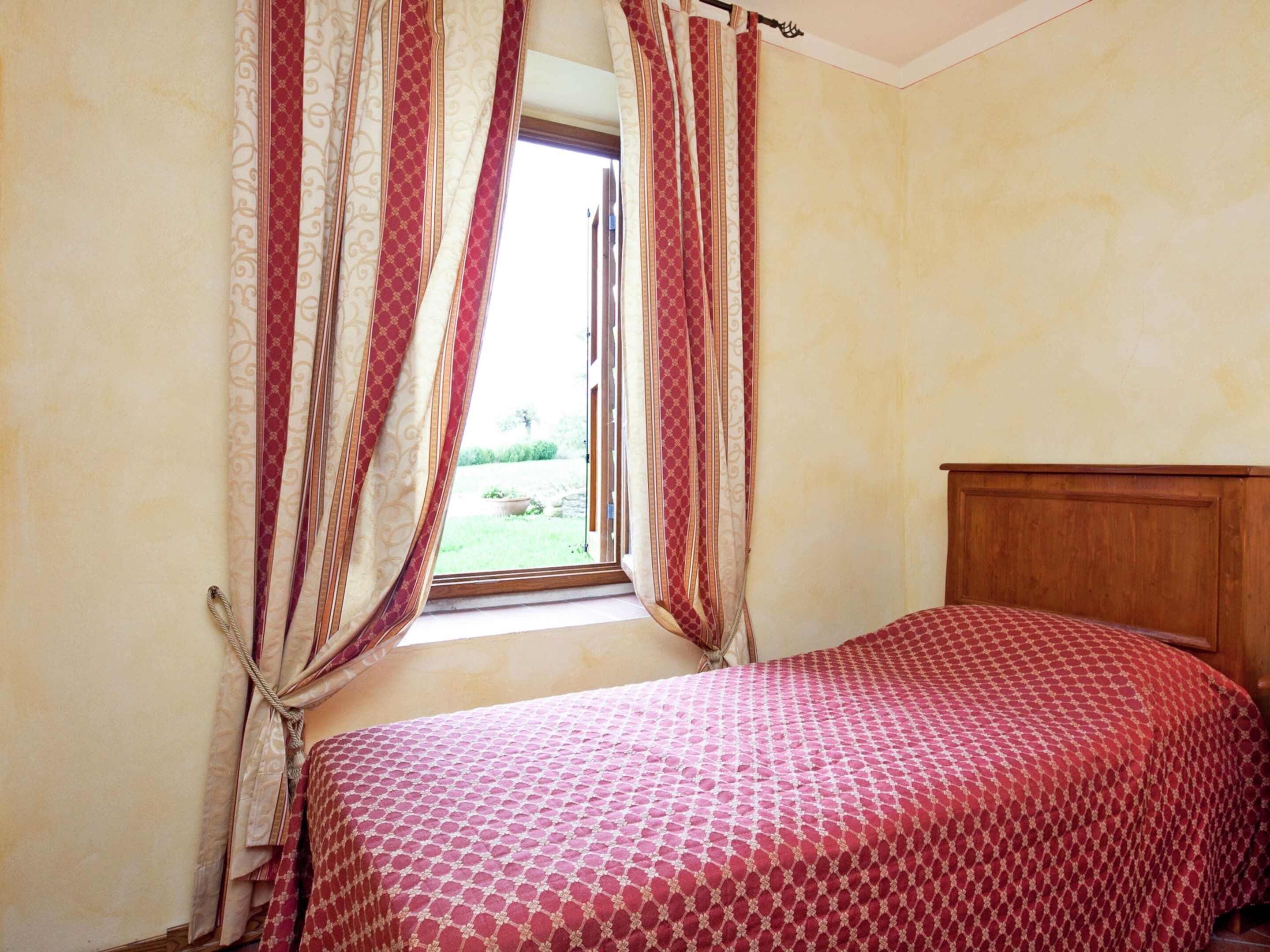 A bedroom with a wooden headboard, a window with red and white curtains, and a patterned bedspread.