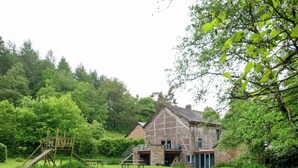 A rustic barn-style house with a stone foundation and a wooden upper structure, surrounded by a lush green lawn and dense trees. - Forest Escape, Luxembourg (Manhay)