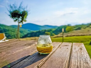 A glass jar with a yellow liquid on a wooden table with a scenic view of green hills and a tree in the background. - Belvilla by OYO Timo (Anghiari)