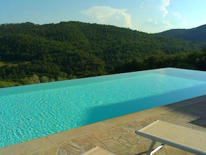 An infinity pool with clear blue water, surrounded by a stone deck and a lounge chair. - Belvilla by OYO Timo (Anghiari)