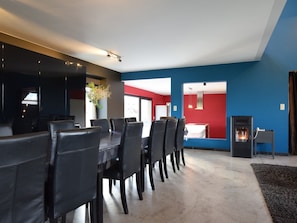 A modern dining area with a long table and chairs, a kitchen in the background, and a fireplace.