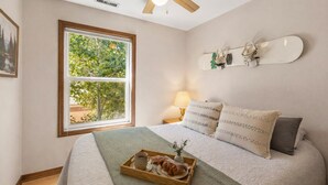 Un dormitorio con un ventanal grande, una cama con una manta verde, almohadas, una mesita de noche con una lámpara y un monopatín en la pared.