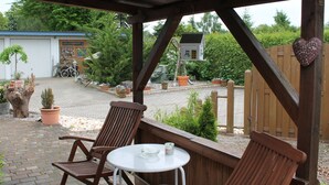 A covered outdoor seating area with two wooden chairs and a small round table, surrounded by potted plants and a wooden fence. - Quaint Holiday Home in Schwaan near Lake (Schwaan)