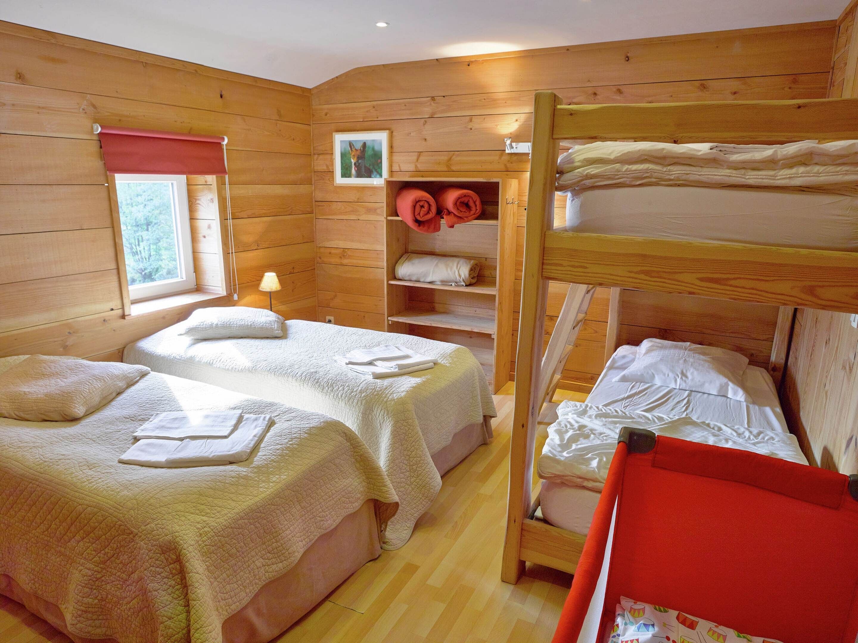 A wooden cabin room with two beds, a bunk bed, a window, and a shelf with towels.