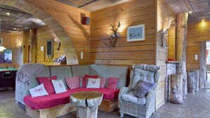 A cozy cabin interior with a wooden ceiling, a sofa with red cushions, a wicker armchair, and antler decorations. - Luxurious Villa in Stoumont With Sauna (Stoumont)