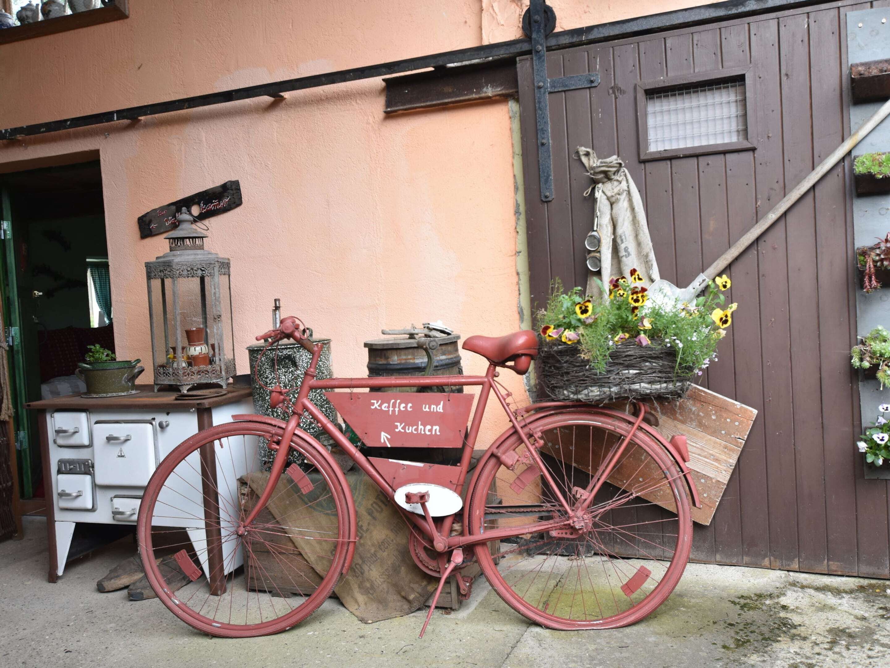 Ein rotes Fahrrad mit einem Blumenkorb steht vor einem Gebäude mit einer braunen Tür und einem Fenster.