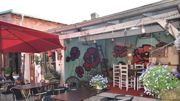 Een terras buiten met houten tafels en stoelen, een rode parasol en een muurschildering met grote bloemen.