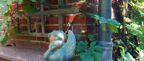 Una estatua de un ángel sobre un alféizar de ladrillo, rodeada de follaje y un poste de metal.