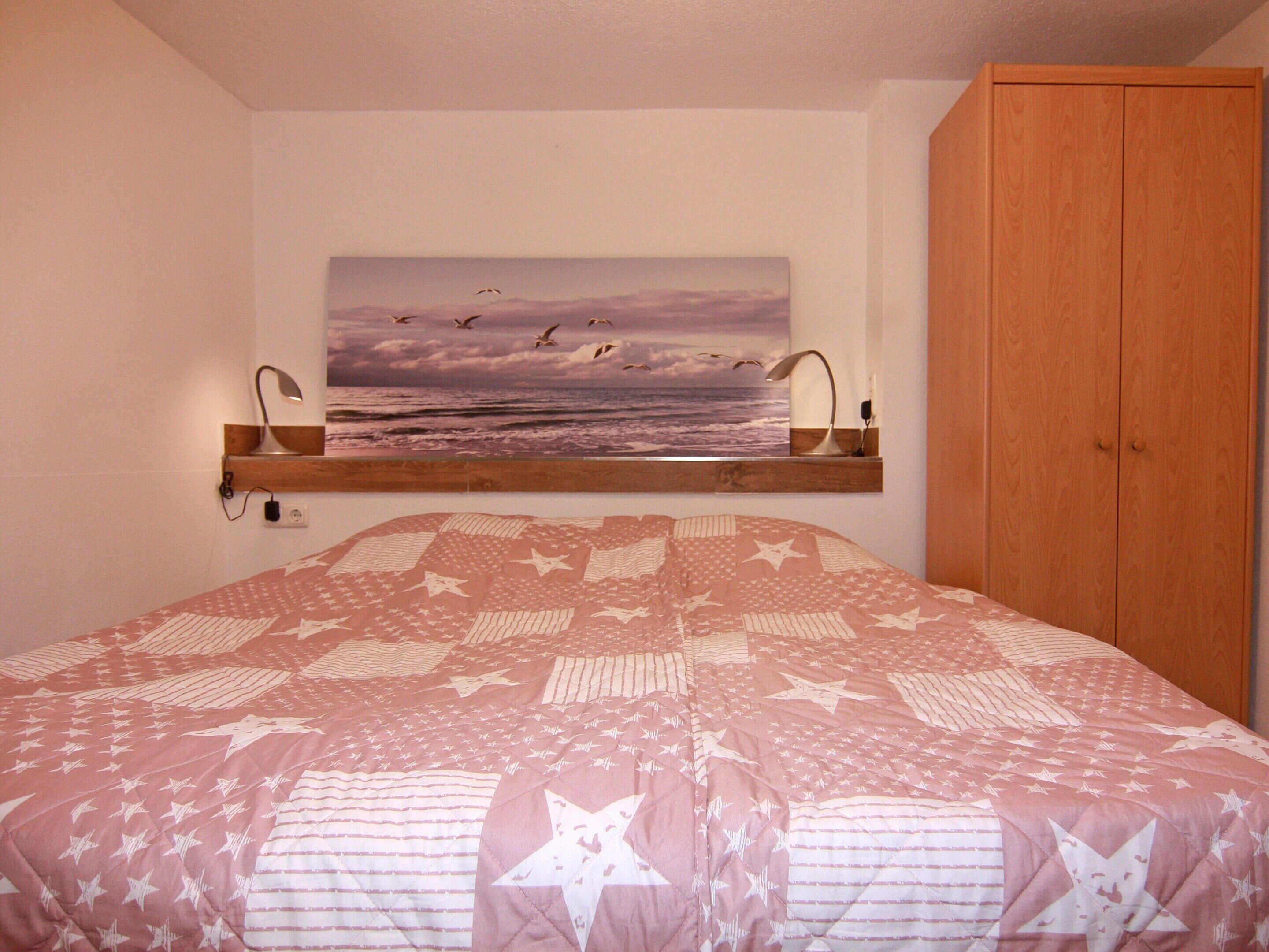 A bedroom with a bed, a wooden wardrobe, a painting of birds flying over the sea, and a shelf with a lamp.