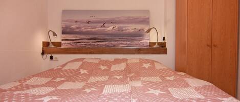 A bedroom with a bed, a wooden wardrobe, a painting of birds flying over the sea, and a shelf with a lamp.