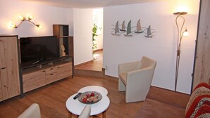 Una sala de estar con televisor, una mesa blanca, un sofá y una silla. En la pared, un cuadro con barcos a vela.