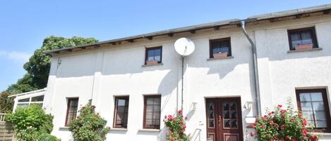 A white building with a brown door, windows, and a satellite dish. There are steps leading to the door, a small table with chairs, and a garden with red flowers.