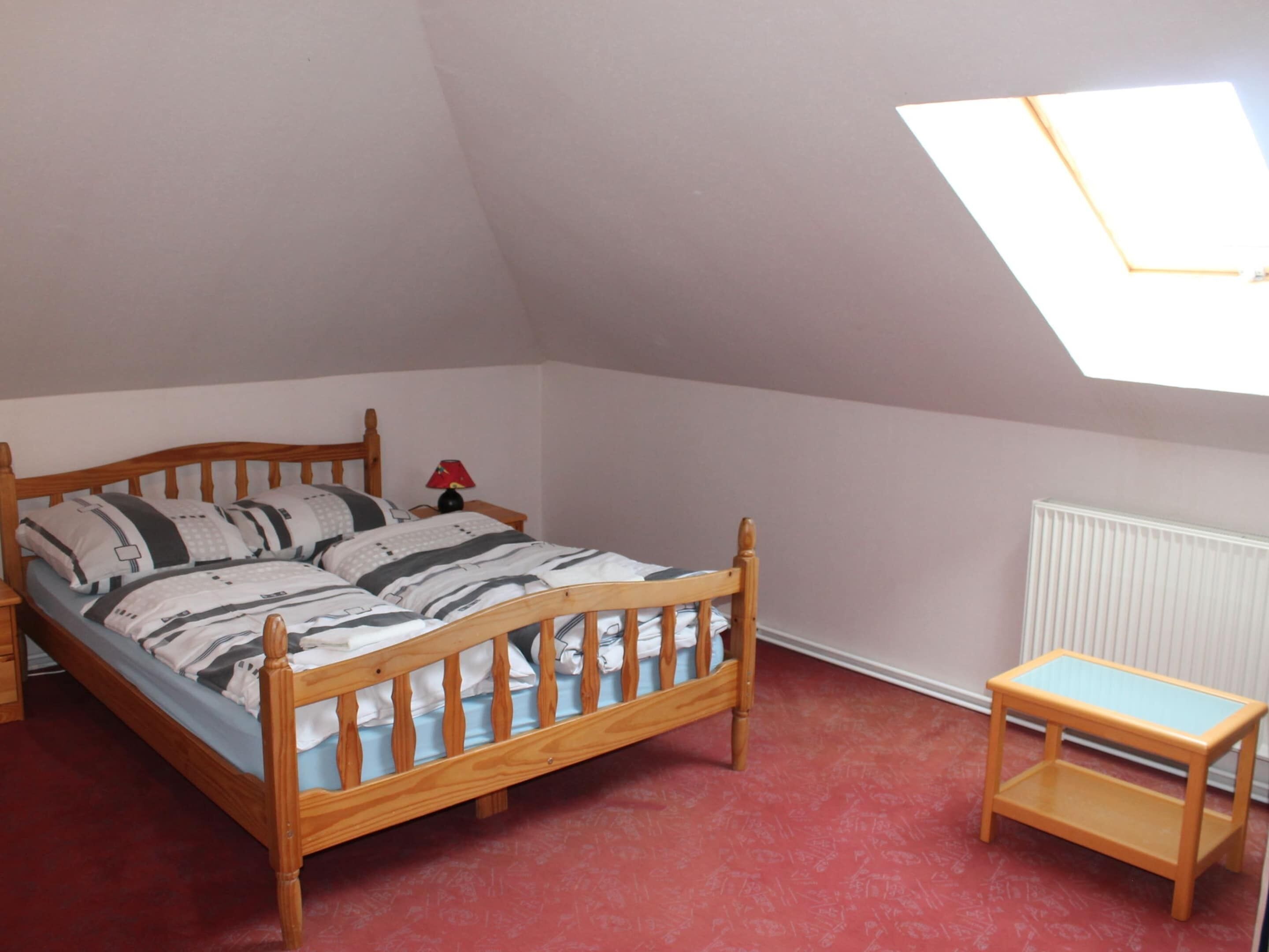 A bedroom with a sloped ceiling, a wooden bed with a striped bedspread, a small table, and a radiator.