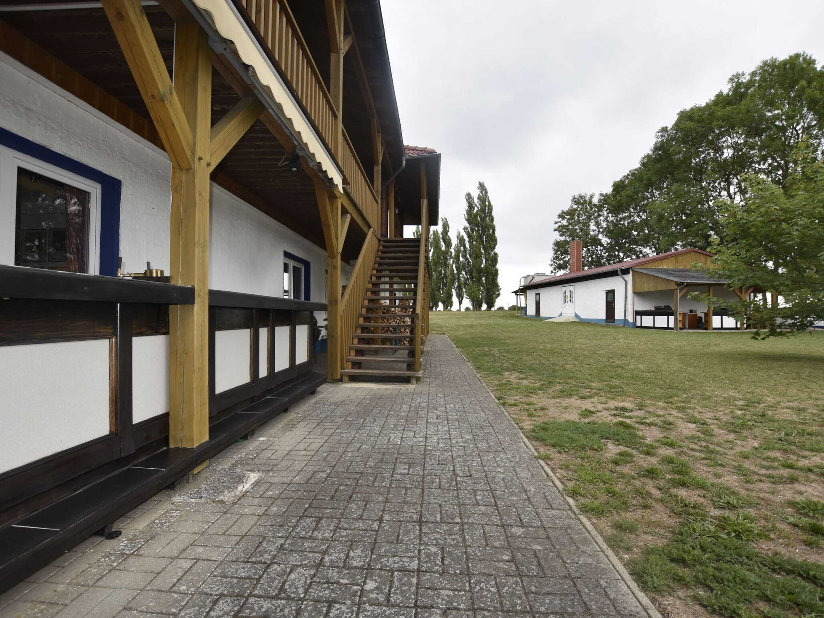 A building with a wooden staircase leading to a balcony, a paved walkway, and a grassy area with trees.