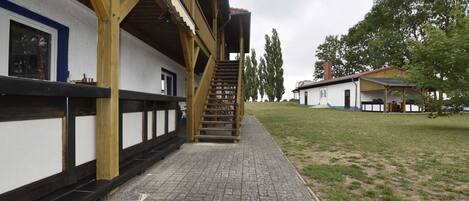 A building with a wooden staircase leading to a balcony, a paved walkway, and a grassy area with trees.