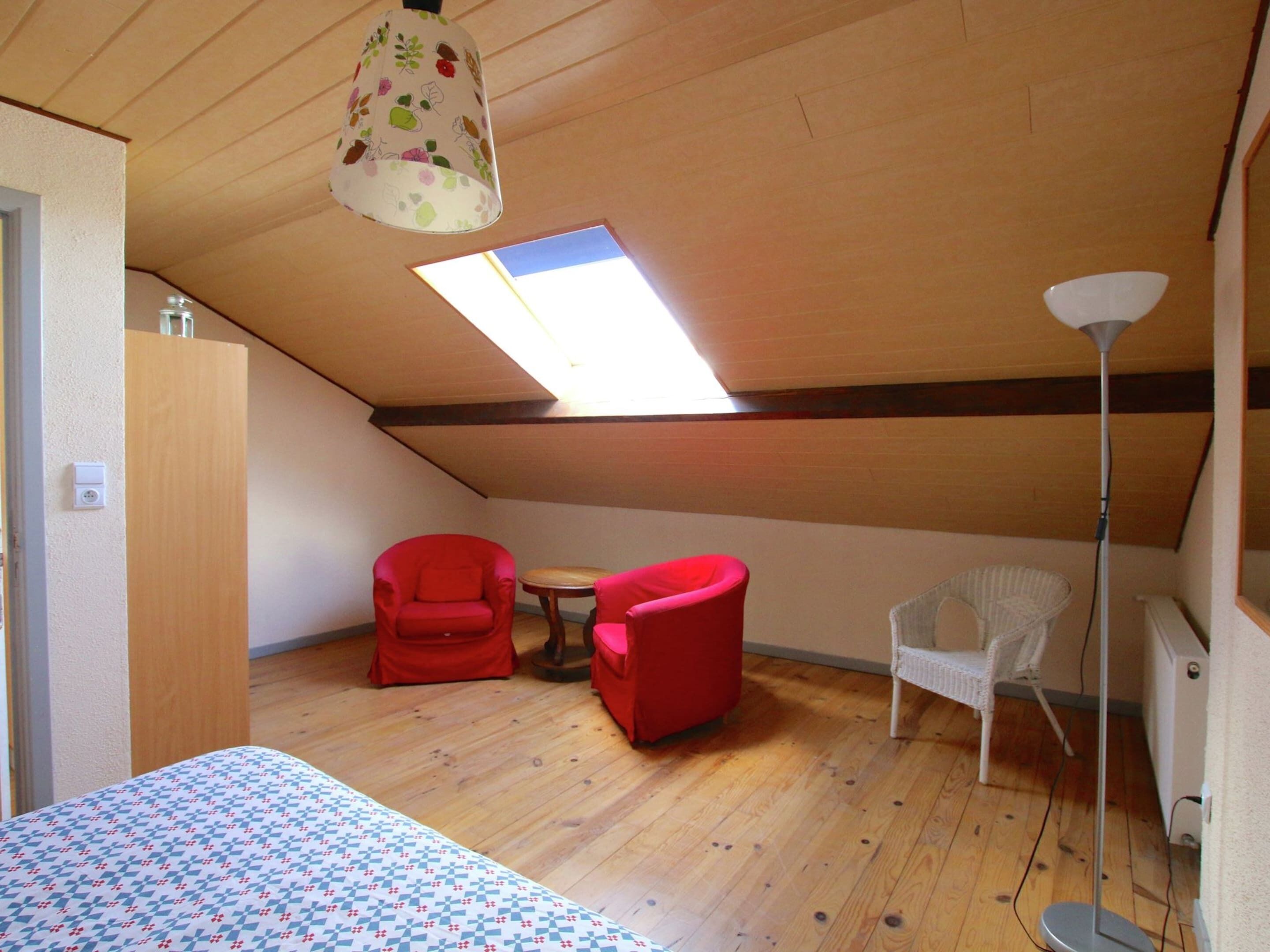 A room with a sloped ceiling, a skylight, a bed with a patterned bedspread, two red chairs, a small round table, and a white chair.