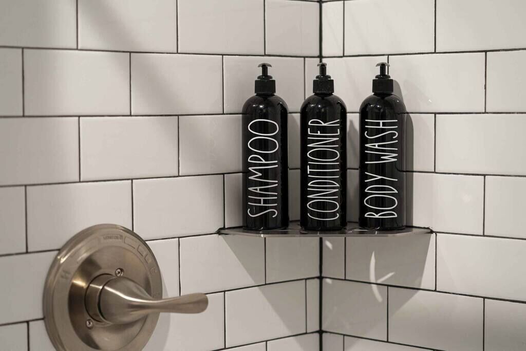 A bathroom with three bottles labeled "SHAMPOO," "CONDITIONER," and "BODY WASH" on a tiled wall.