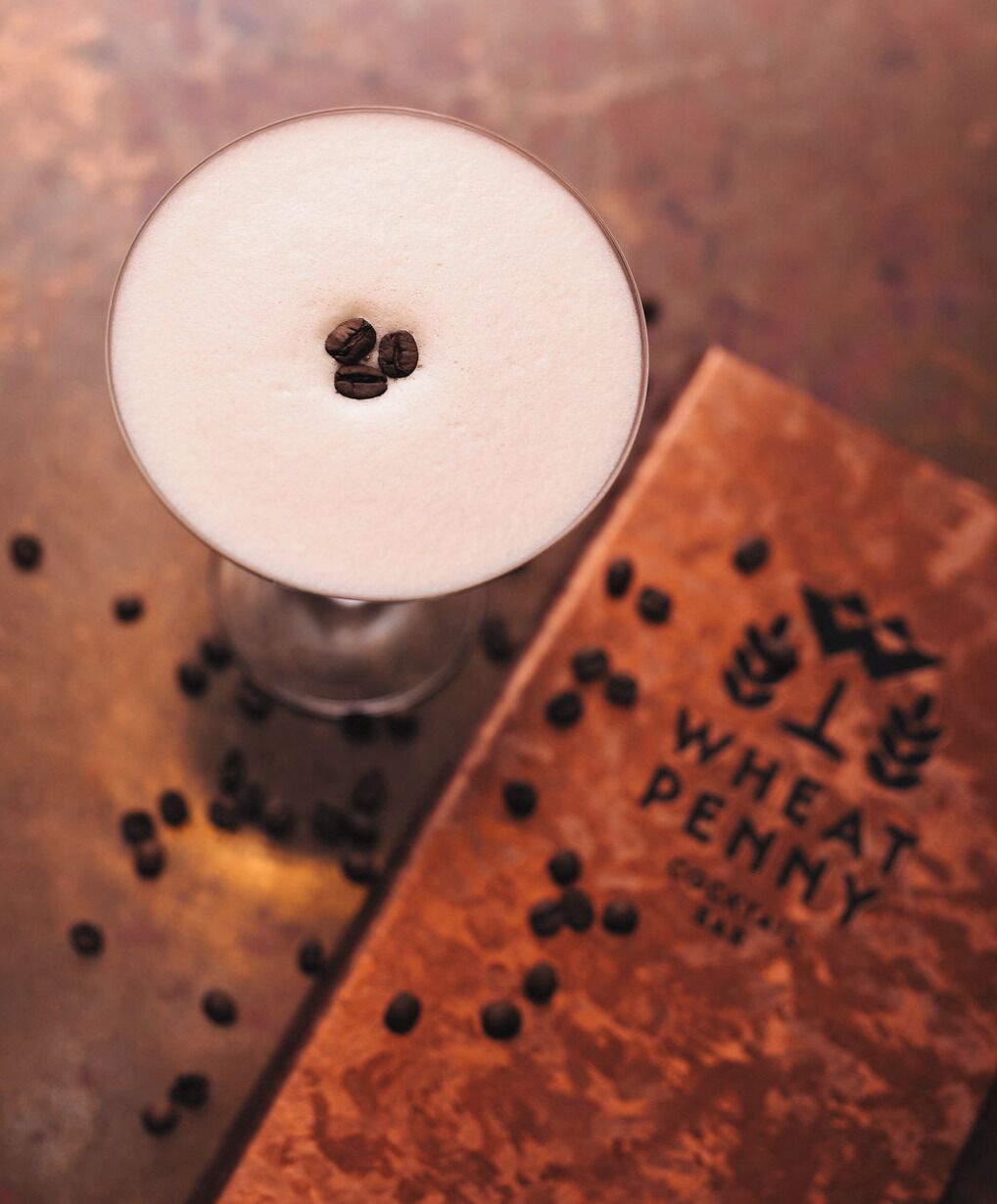 Un verre contenant une boisson claire, surmonté de grains de café, posé sur une surface en bois parsemée de grains de café et à côté d’une boîte étiquetée « WHEAT PENNY COCKTAIL ».