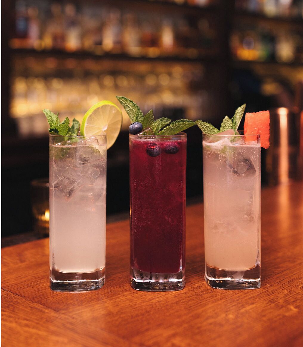 Three tall glasses of iced drinks on a wooden bar counter.