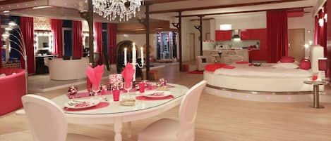 A modern interior with a round dining table, pink chairs, a chandelier, and a kitchen area.