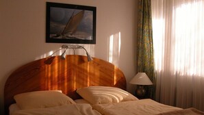 Una habitación de hotel con cabecera de madera, cama con ropa blanca, un cuadro enmarcado de un velero, una lámpara y cortinas traslúcidas.