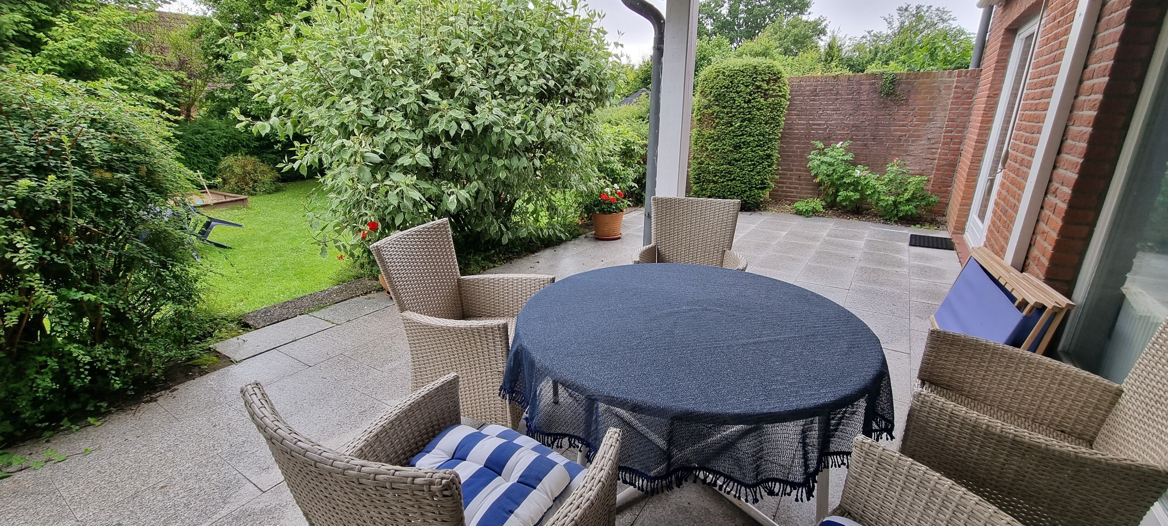 Un patio con mobiliario de mimbre y una mesa redonda, rodeado de abundante vegetación y un muro de ladrillo.