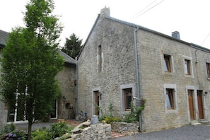 A stone building with a sloped roof, multiple windows, and a small garden area with plants and a tree. - Holiday Home in Longueville (Durbuy)