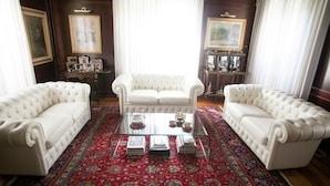 A room with three white leather sofas arranged around a glass coffee table with books and decorative items. The room features dark wooden walls, ornate paintings, and a richly patterned red carpet. - Elegant Art Nouveau Villa With Private Pool Near the Lake (Desenzano del Garda)