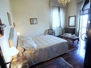 A bedroom with a large bed, a chair, a table with books, and a chandelier. - Elegant Art Nouveau Villa With Private Pool Near the Lake (Desenzano del Garda)
